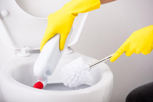 Female hand in yellow rubber glove is cleaning toilet bowl using brush.