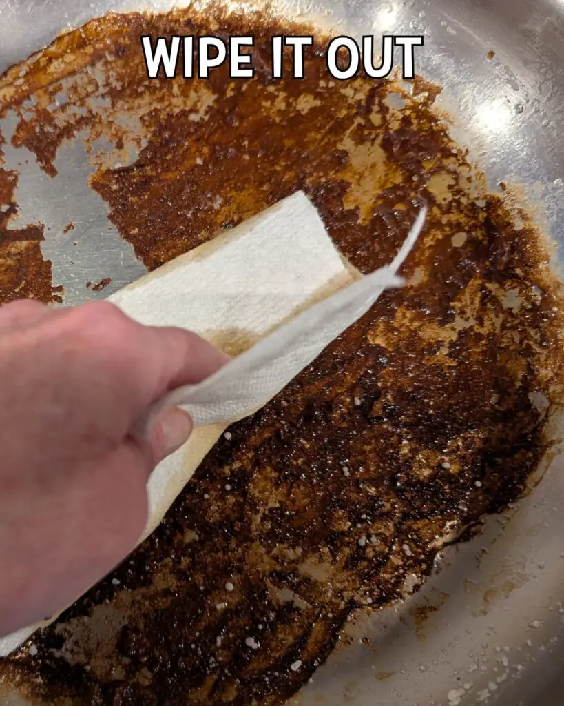 A hand holding a paper towel is wiping out a heavily burnt pan, illustrating how to clean a burnt pan.