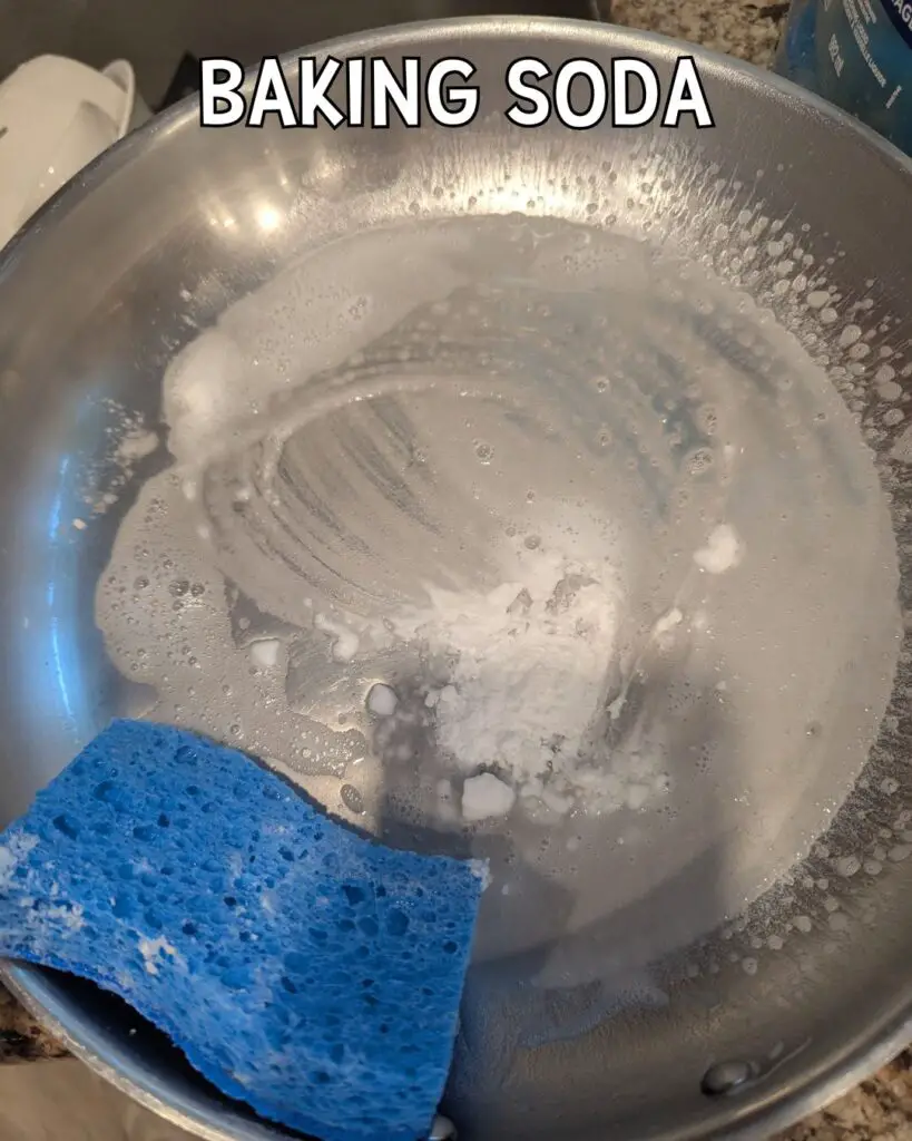 A stainless steel pan with a layer of soapy water and baking soda, accompanied by a blue sponge, is shown on a countertop.
