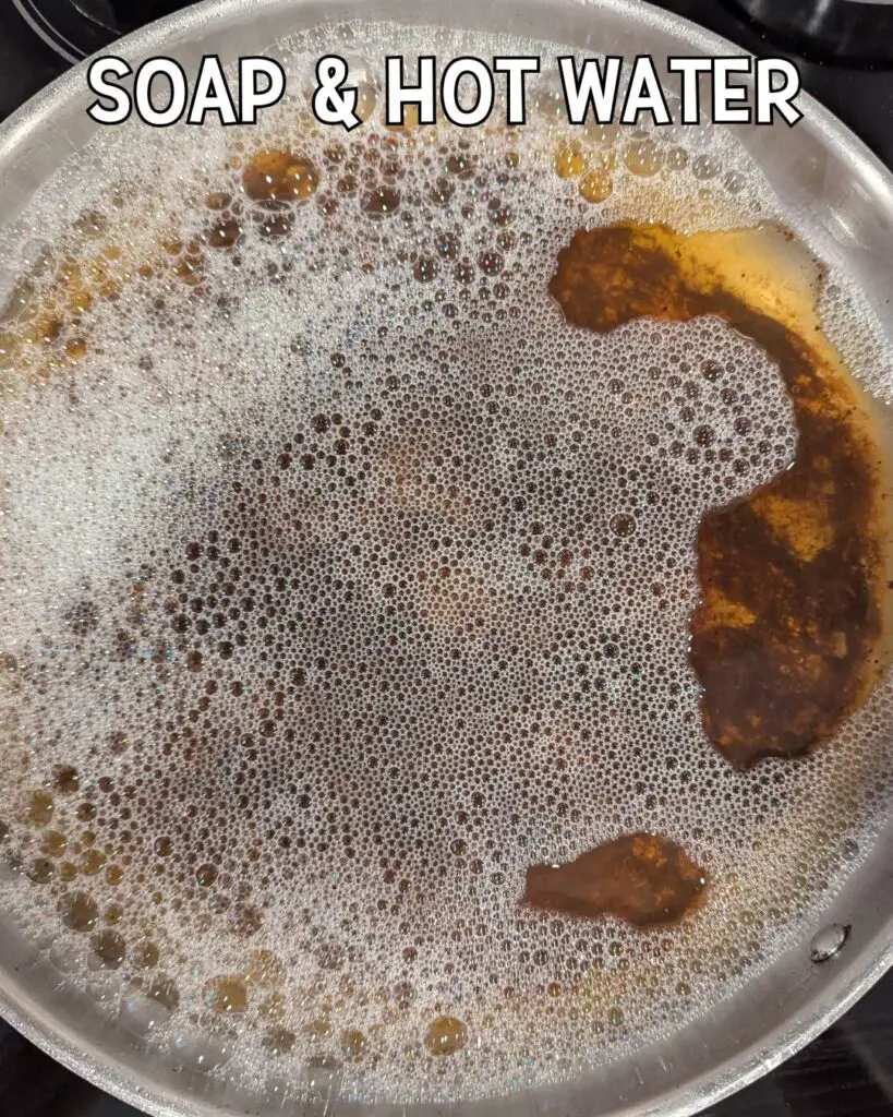 A stainless steel pan filled with soapy, foamy water and remnants of burnt food, illustrating a method for cleaning a burnt stainless steel pan.